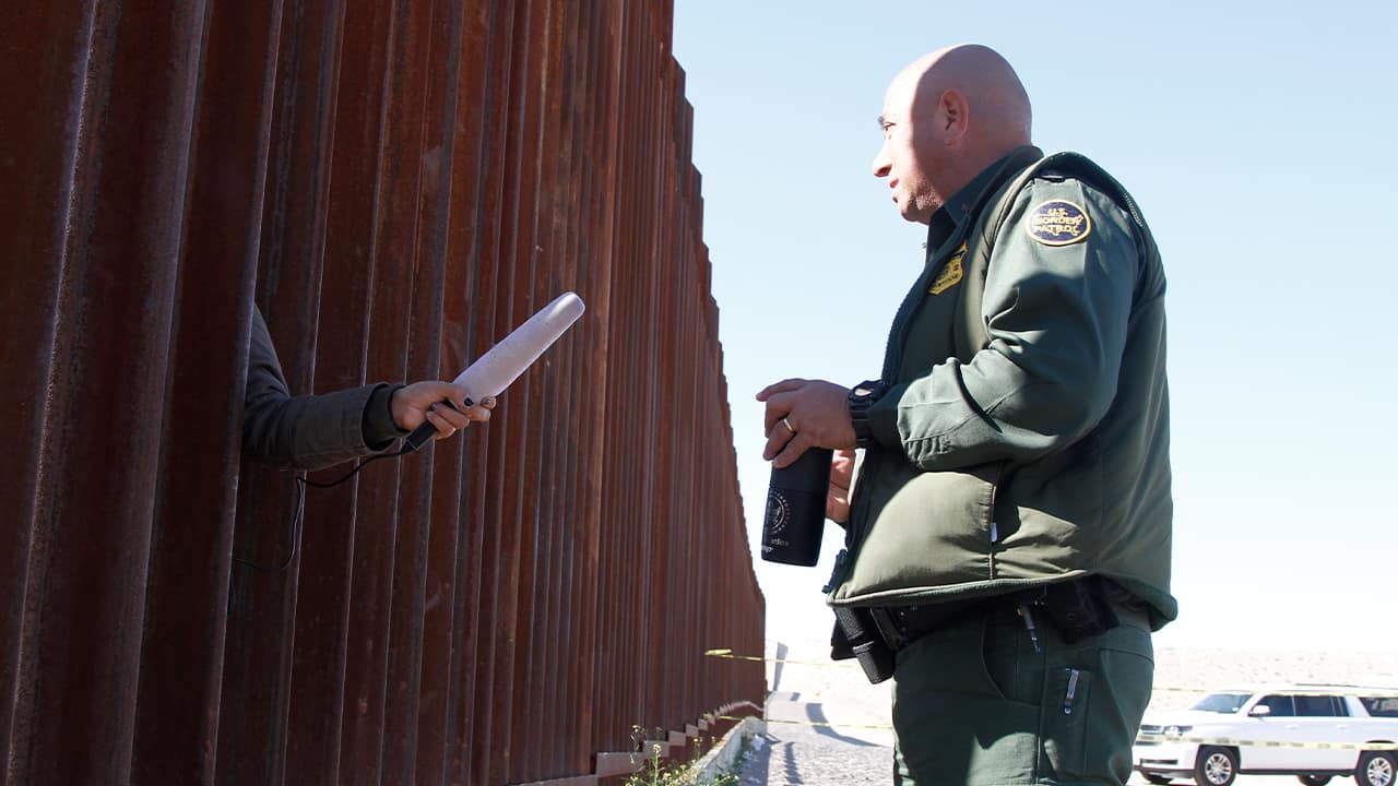 Photo of a journalist sticking her microphone through a through a border fence to interview a Border Patrol agent