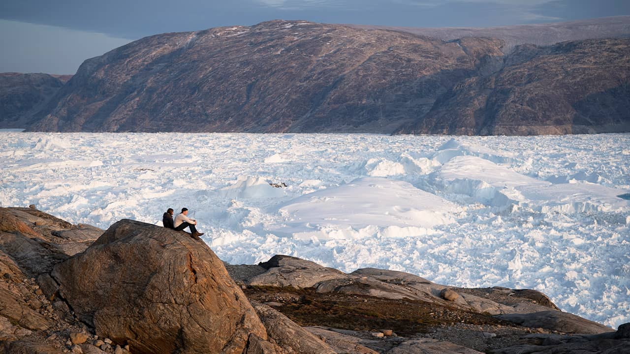 Photo of researchers sitting on a rock overlooking the Helheim glacier in Greenland