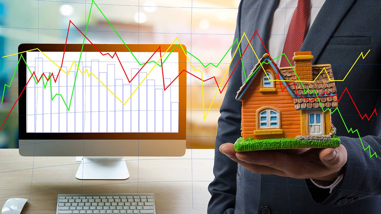 Photo of man holding a house next to a computer with stock charts