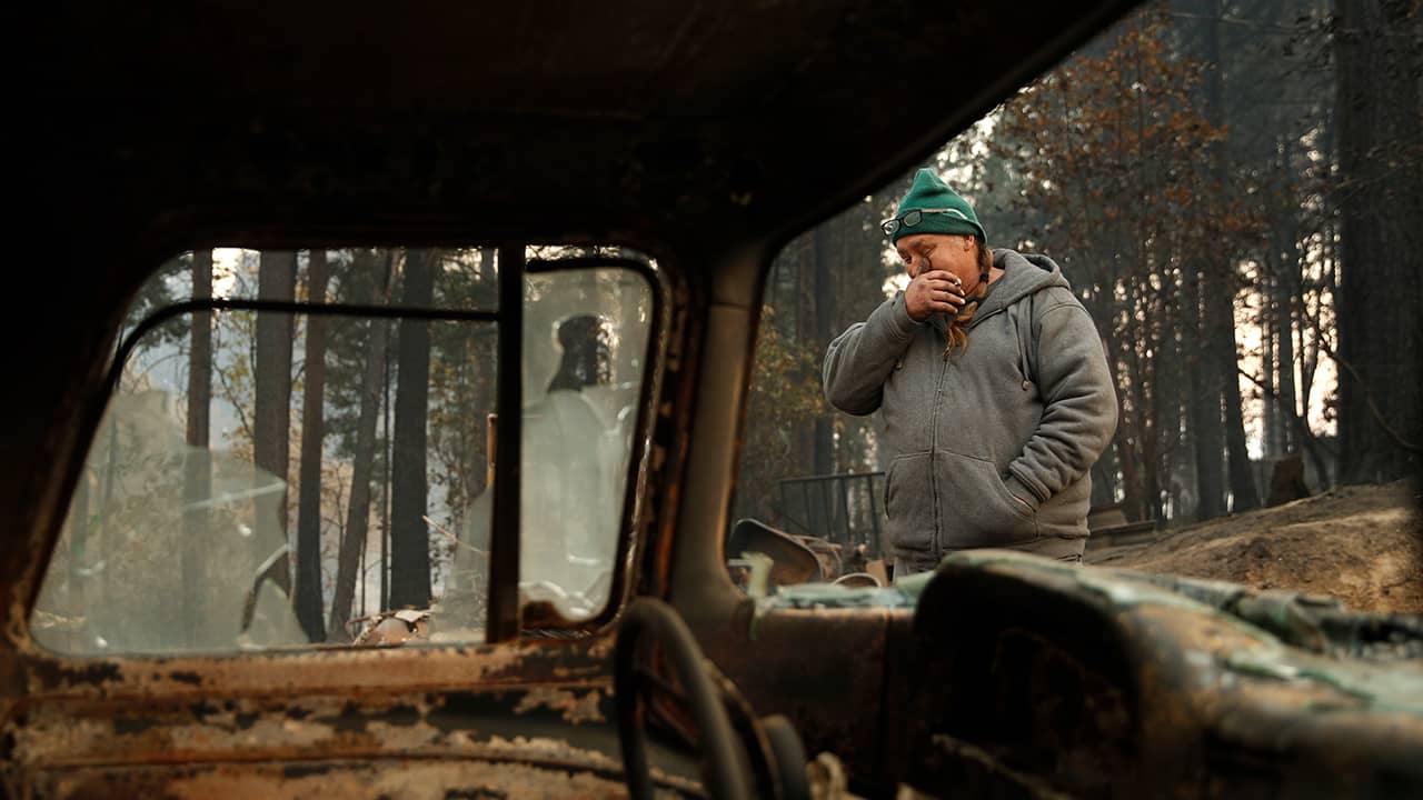 Photo of a wildfire victim wiping his eyes as he walks by a burned car