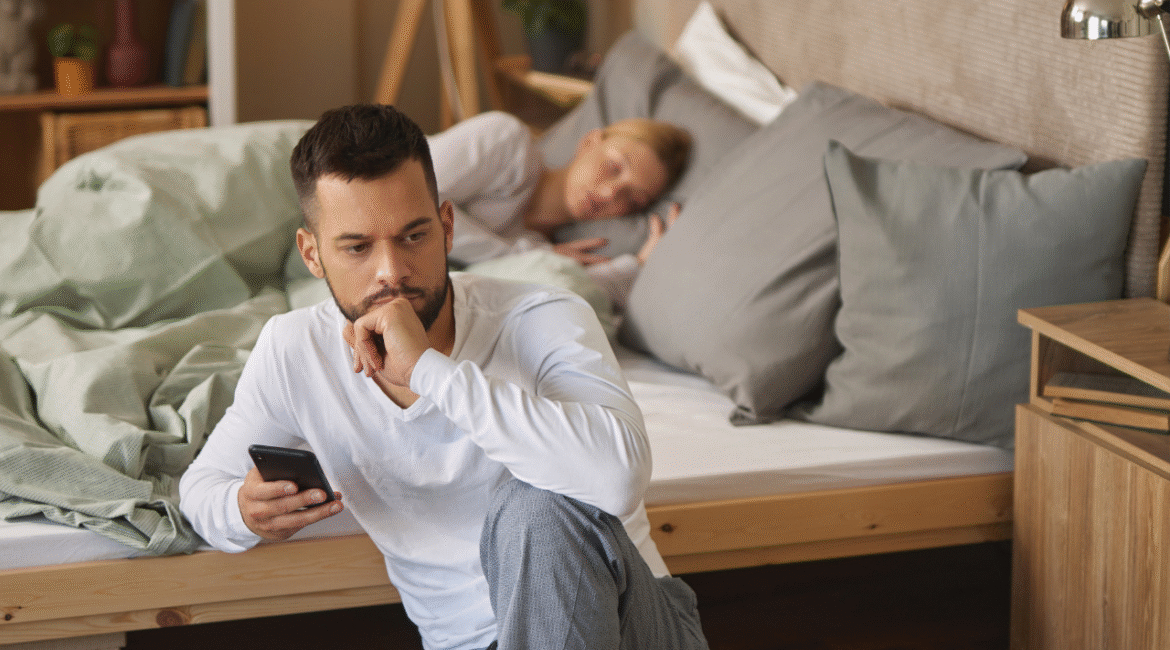 Man with smart phone in bedroom, woman sleeping in the background