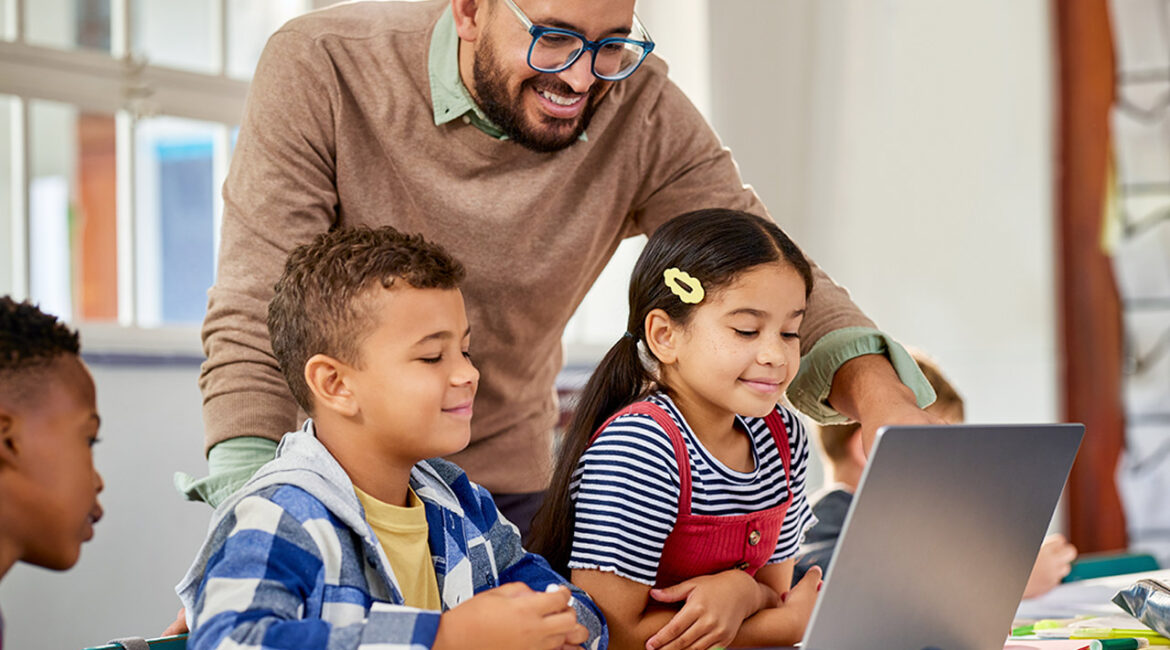 Happy smiling teacher and elementary students using laptop in class during technology lesson. Primary school educator giving a digital literacy lesson in an elementary school with computer.