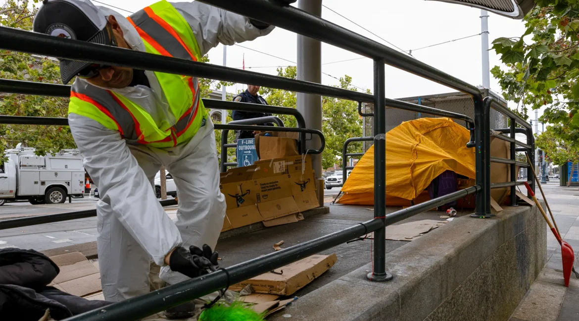 City worker in protective gear removes belongings near a yellow tent during a homeless encampment cleanup at an urban transit stop.
