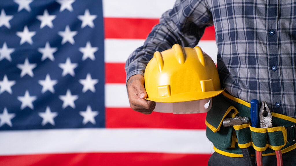 Close up shot of labour with tools or equipment by holding helmet in hand in front of USA flag - concept of maintenance service, labour day and employment.