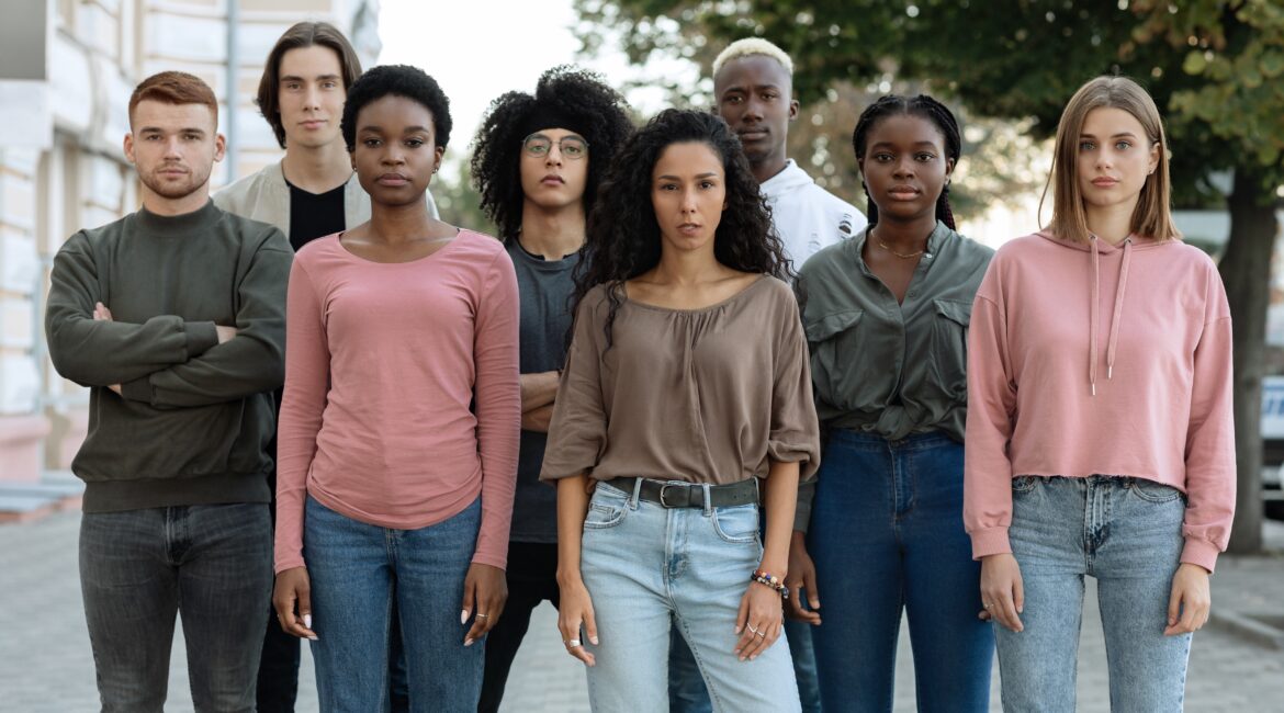 A group of young adults of different racial and ethnic backgrounds stand side by side outdoors, looking directly at the camera with serious expressions.