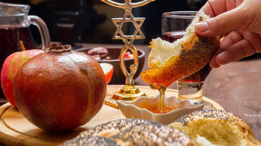 Women's hands dip challah in honey at a table with traditional wine and pomegranate on Rosh Hashanah.