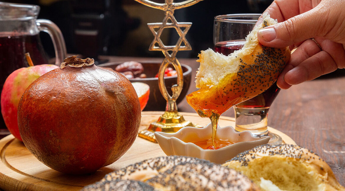 Women's hands dip challah in honey at a table with traditional wine and pomegranate on Rosh Hashanah.
