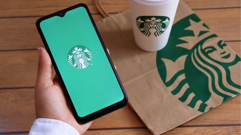 Asset id: 1874117719 Bahia, Brazil - December 11, 2020. Woman holding smartphone with Starbucks app on screen, and Starbucks cup and paper bag in the background
