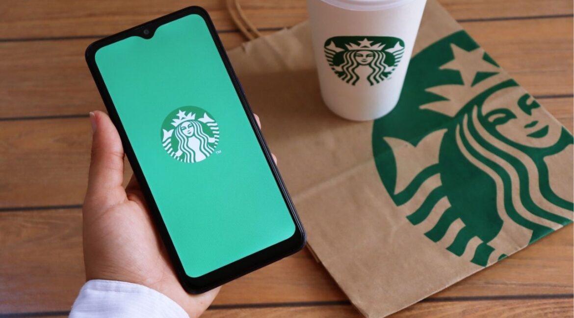 Asset id: 1874117719 Bahia, Brazil - December 11, 2020. Woman holding smartphone with Starbucks app on screen, and Starbucks cup and paper bag in the background