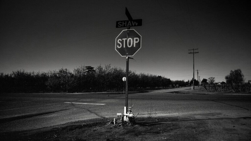 Black and white photo illustration of a deadly Fresno County intersection, its stop sign, and a white cross memorial with flowers there