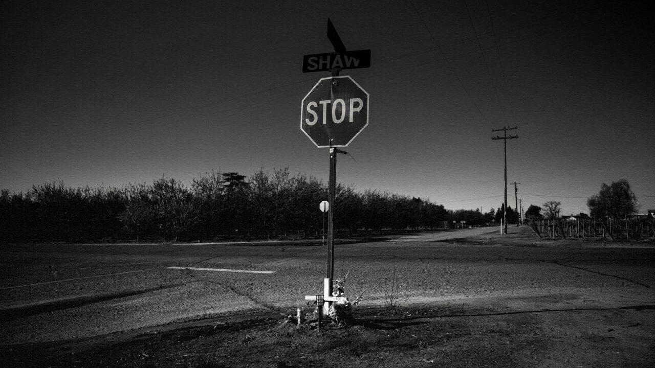Black and white photo illustration of a deadly Fresno County intersection, its stop sign, and a white cross memorial with flowers there
