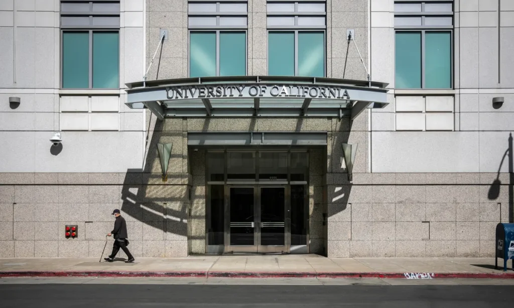 Image of a person with a cane walking past the entrance of the imposing University of California Office of the President building in Oakland on a spring day.