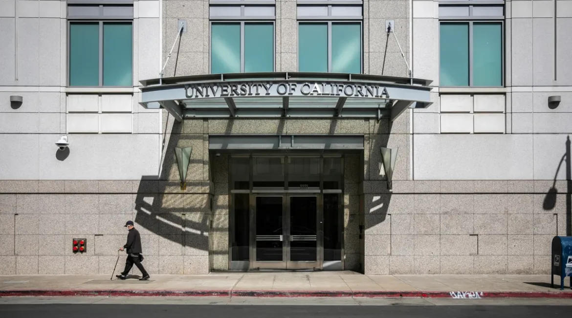 Image of a person with a cane walking past the entrance of the imposing University of California Office of the President building in Oakland on a spring day.