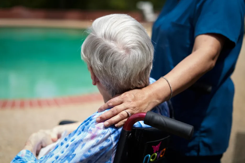 Image of a caregiver, in a dark blue smock, with her hand on the shoulder of an elderly client who is sitting in a wheelchair.