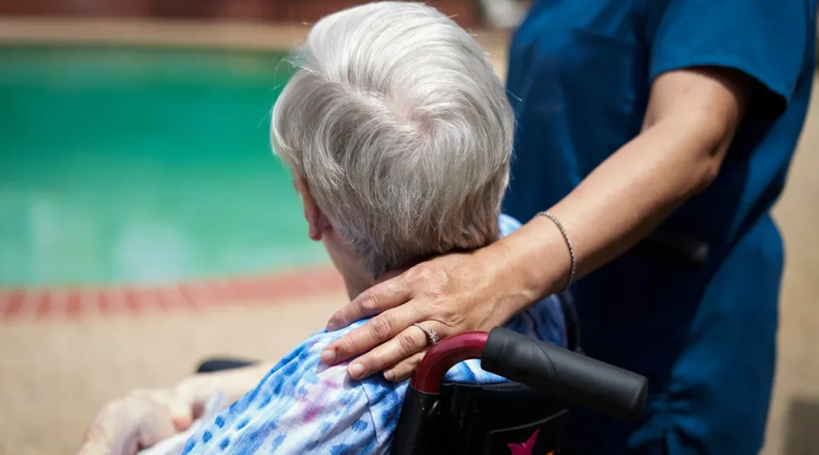 Image of a caregiver, in a dark blue smock, with her hand on the shoulder of an elderly client who is sitting in a wheelchair.