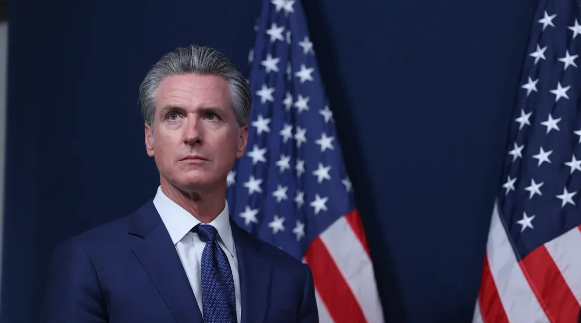 Image of Gov. Newsom, in a blue suit with a white shirt and blue tie, standing in front of American flags
