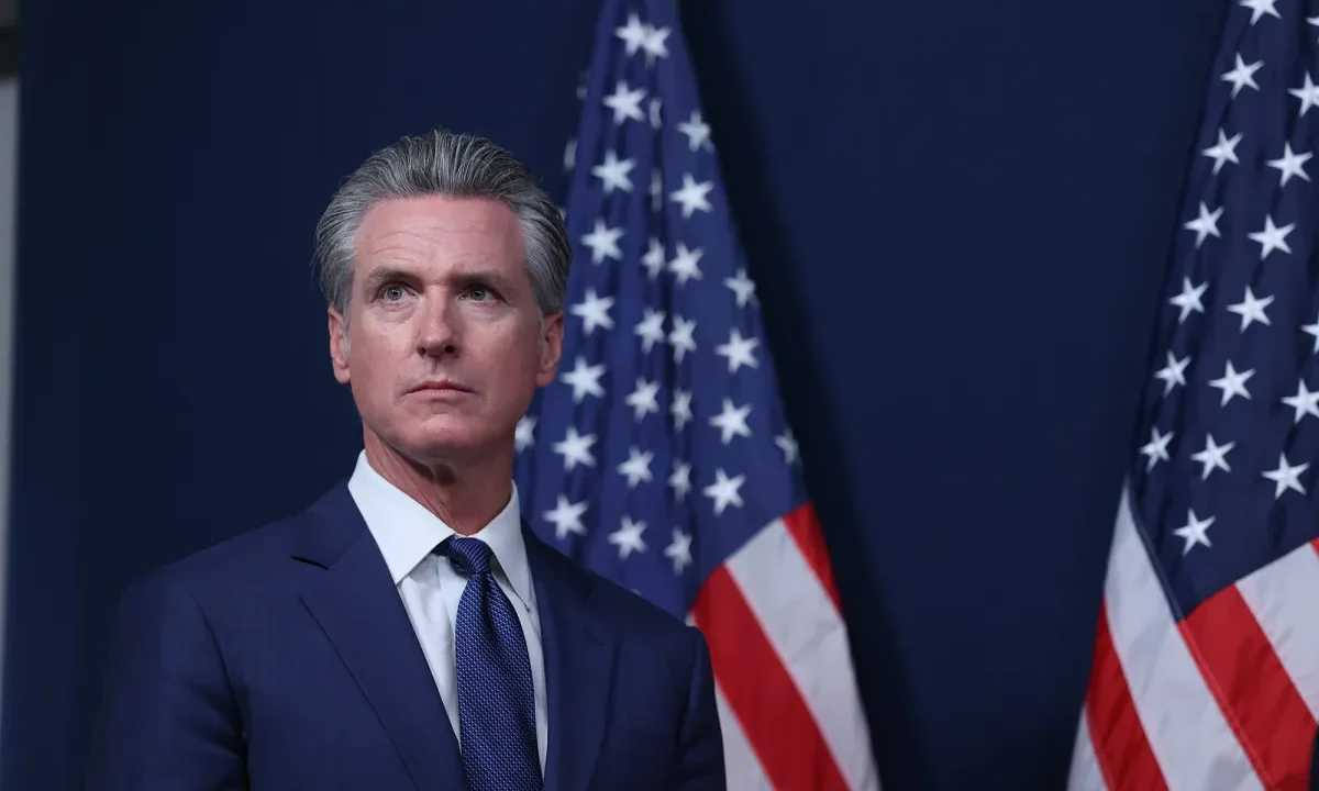 Image of Gov. Newsom, in a blue suit with a white shirt and blue tie, standing in front of American flags