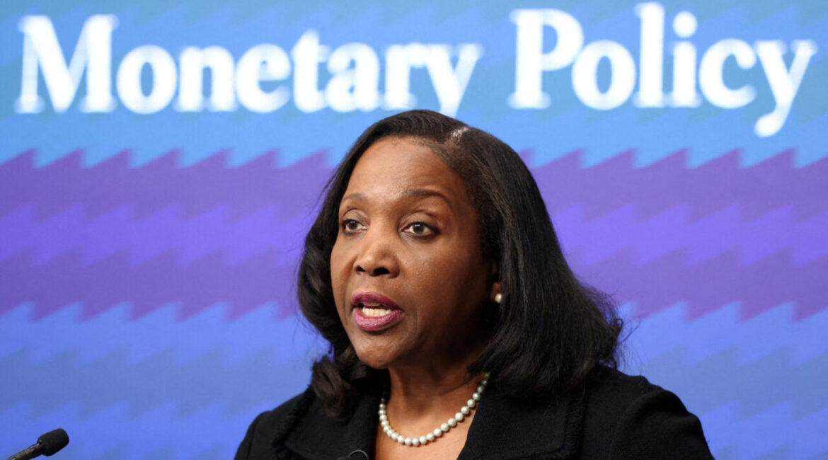 Image of Federal Reserve Governor Lisa Cook in a black dress with a white necklace in front of a blue and purple sign that says "Monetary Policy"