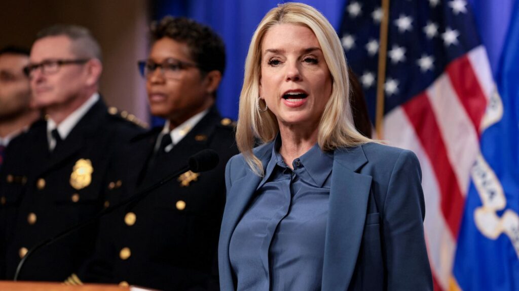 Image of US Attorney General Pam Bondi, wearing a light blue suit, speaking and standing in front of an American Flag at a news conference. There are uniformed officers behind her.
