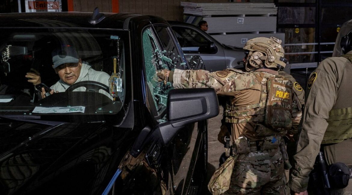 Image of camo-clad federal immigration agent smashing the window of a car with a man inside