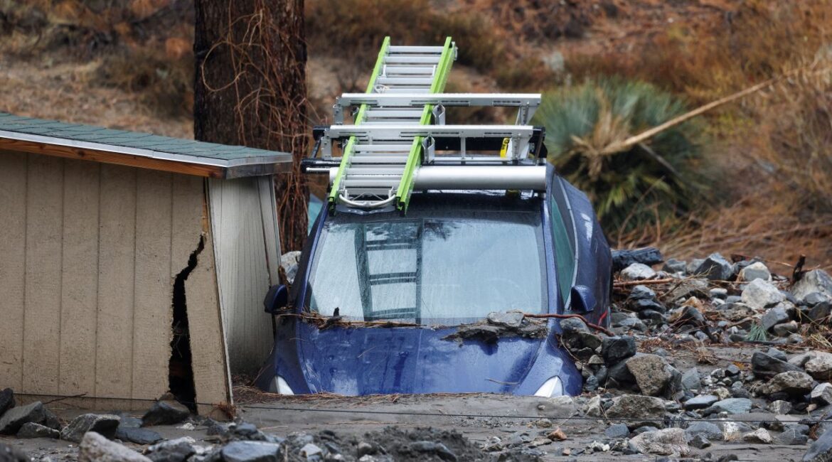 Image of a blue car buried nose first in mud next to a storage shed and under a tree, The care has a rack, which is holding a ladder, on top.
