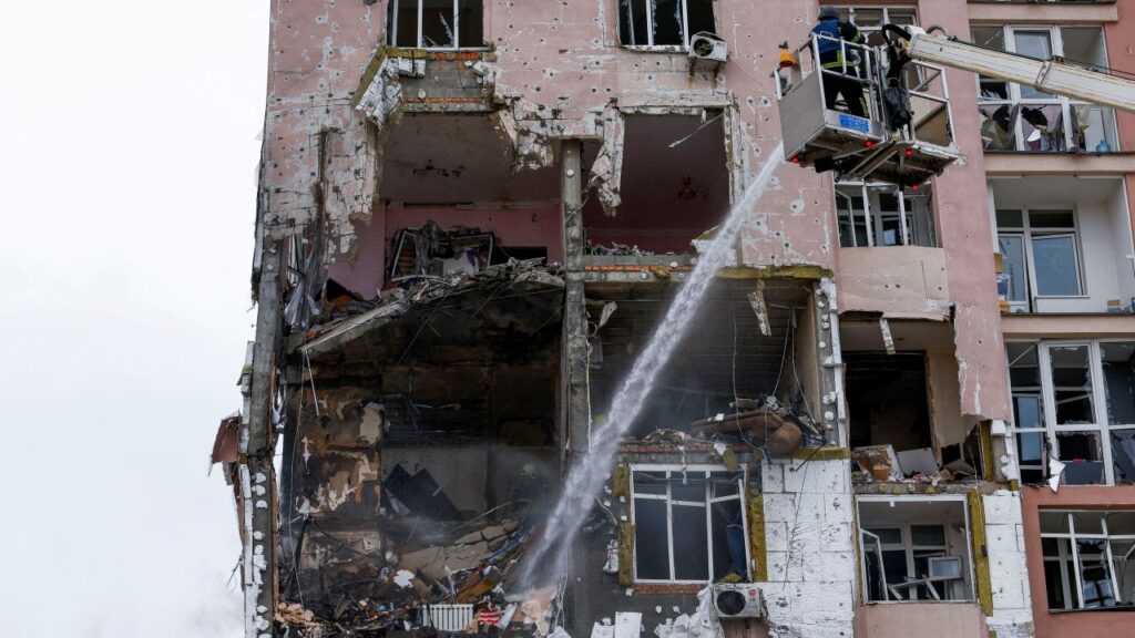 Image of a firefighter on an elevated lift dousing embers at a Ukrainian apartment building hit by Russian drones.