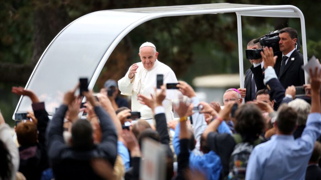 Image of Pope Francis blessing the crowd, which is raising cell phones to video the pontiff.
