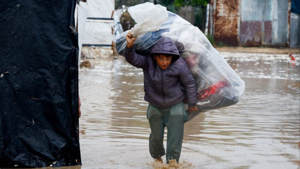 A displaced Palestinian carries belongings in a flooded tent camp on a rainy day in Nuseirat, central Gaza Strip, December 12, 2025. (Reuters/Mahmoud Issa)