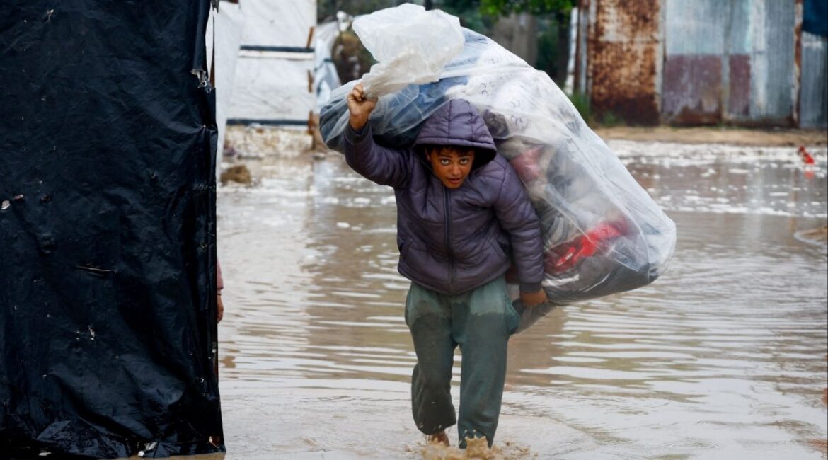 A displaced Palestinian carries belongings in a flooded tent camp on a rainy day in Nuseirat, central Gaza Strip, December 12, 2025. (Reuters/Mahmoud Issa)