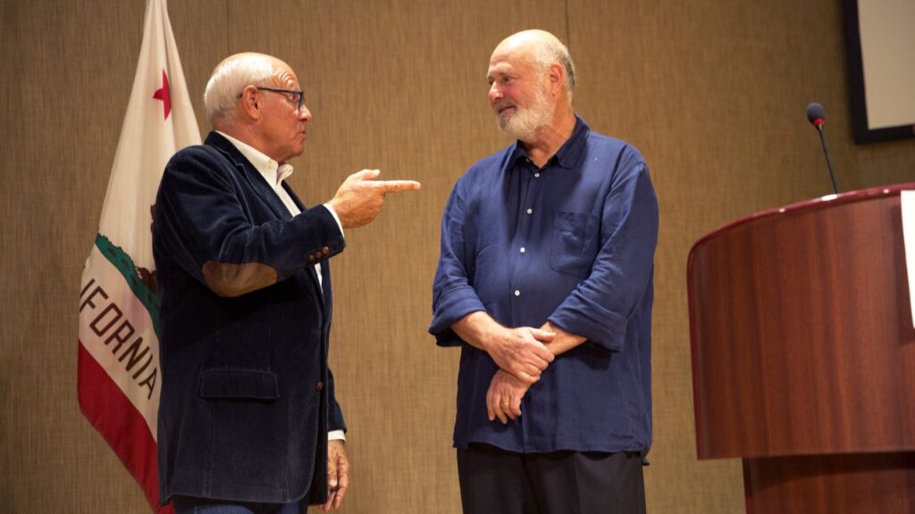 Image of then LA police commissioner Steve Soboroff, in a corduroy coat with elbow patches, talking to Rob Reiner, who is wearing dark baggy pants and a loose blue shirt