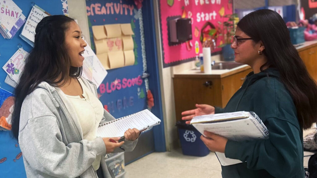 A Clovis High CTE student in a white blouse and gray sweater talks to her teacher in a dark outfit. Both are holding notebooks