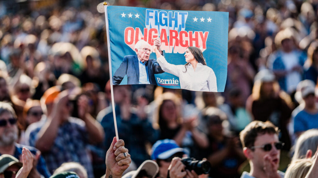 Image of a large protest with a blue hand flag flag raised high stating "Fight Oligarchy." The flag features pictures of Sen. Bernie Sanders and Rep. Alexandria Ocasio-Cortez.