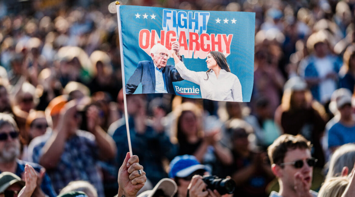 Image of a large protest with a blue hand flag flag raised high stating "Fight Oligarchy." The flag features pictures of Sen. Bernie Sanders and Rep. Alexandria Ocasio-Cortez.