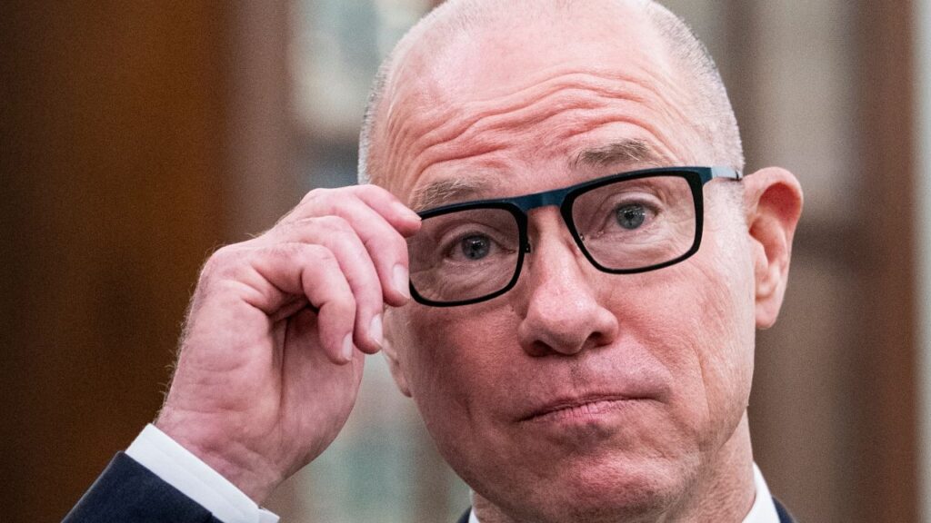 Image of Bryan Bedford. He's wearing a dark blue suit, white shirt, and yellow tie, and tugging at his eyeglasses.