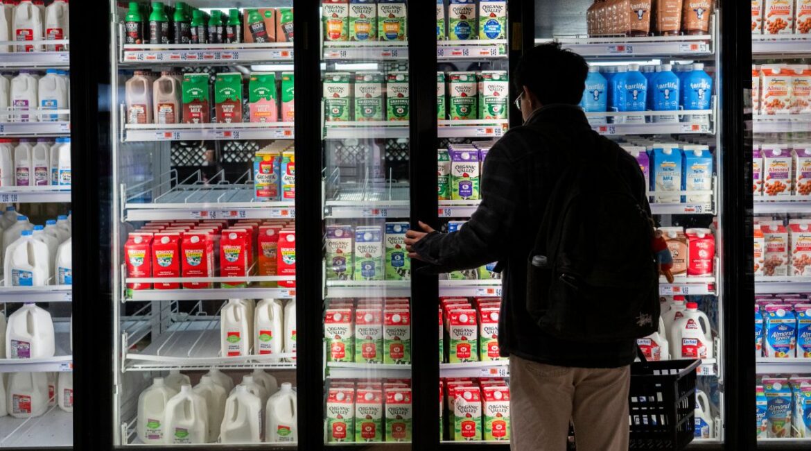 Image of a shopper in a dark coat and tan pants opening the dairy section door at a grocery store