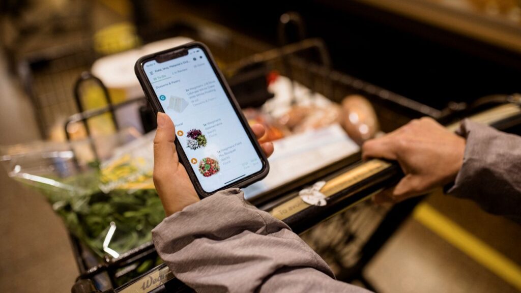 Image of hands pushing a grocery cart while checking prices on a smart phone