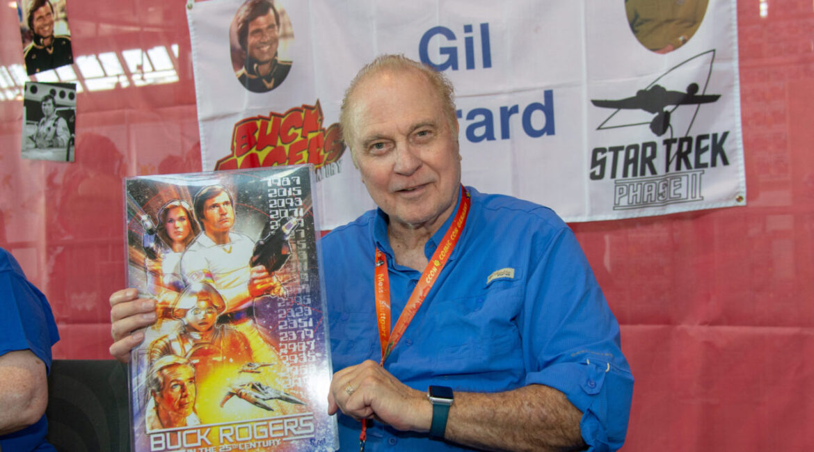 Image of Gil Gerard in a blue shirt holding up a Buck Rogers poster