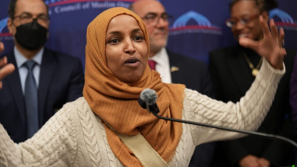 Image of Rep. Ilhan Omar speaking at the Capitol in Washington, D.C. She's wearing a oak-colored knit sweater with a caramel colored hijab
