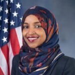 Congressional portrait of Ilman Omar standing in front of an American flag and wearing a dark blazer and multi-colored hijab