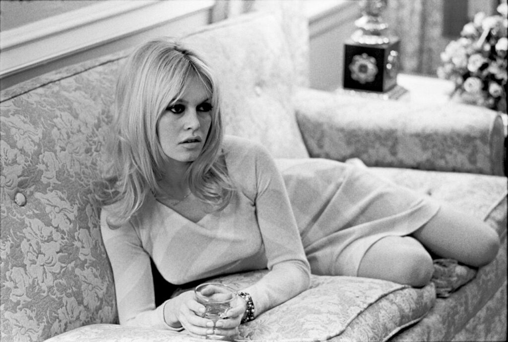 Black and white image of Brigitte Bardot dressed in while posing for the camera on a couch with a drink in her hand.