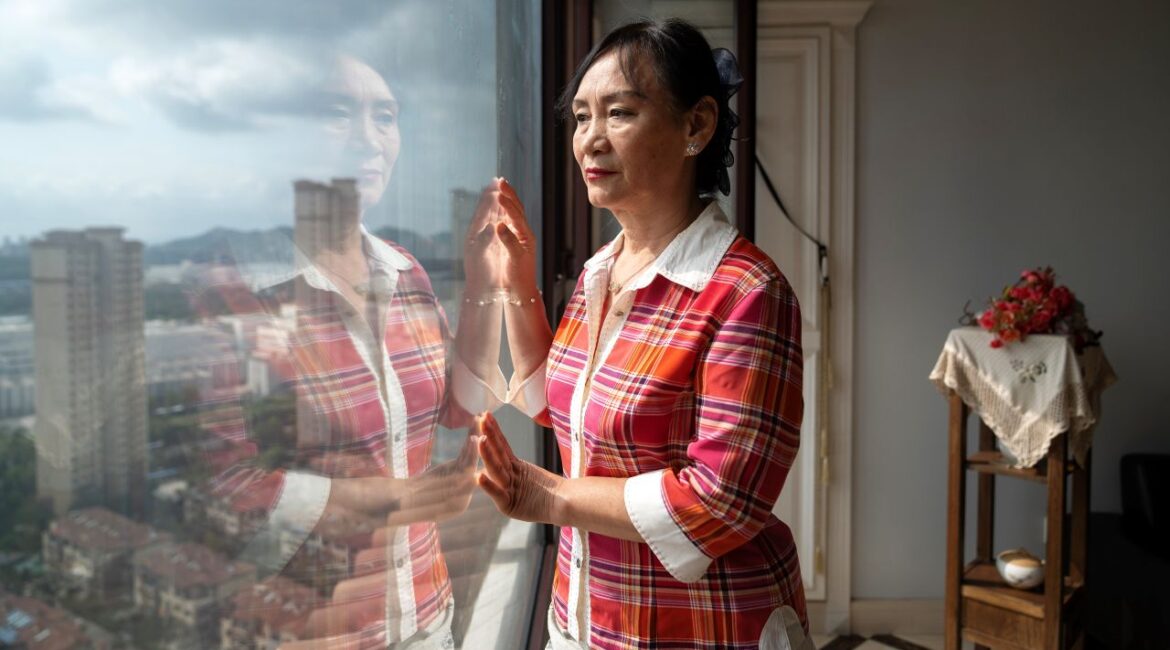 Angel Zhao, Dr. Yan’s mother, at her home in Qingdao, China, peering out the window of her apartment. She is wearing a red-hue tartan shirt-jacket over a white shirt