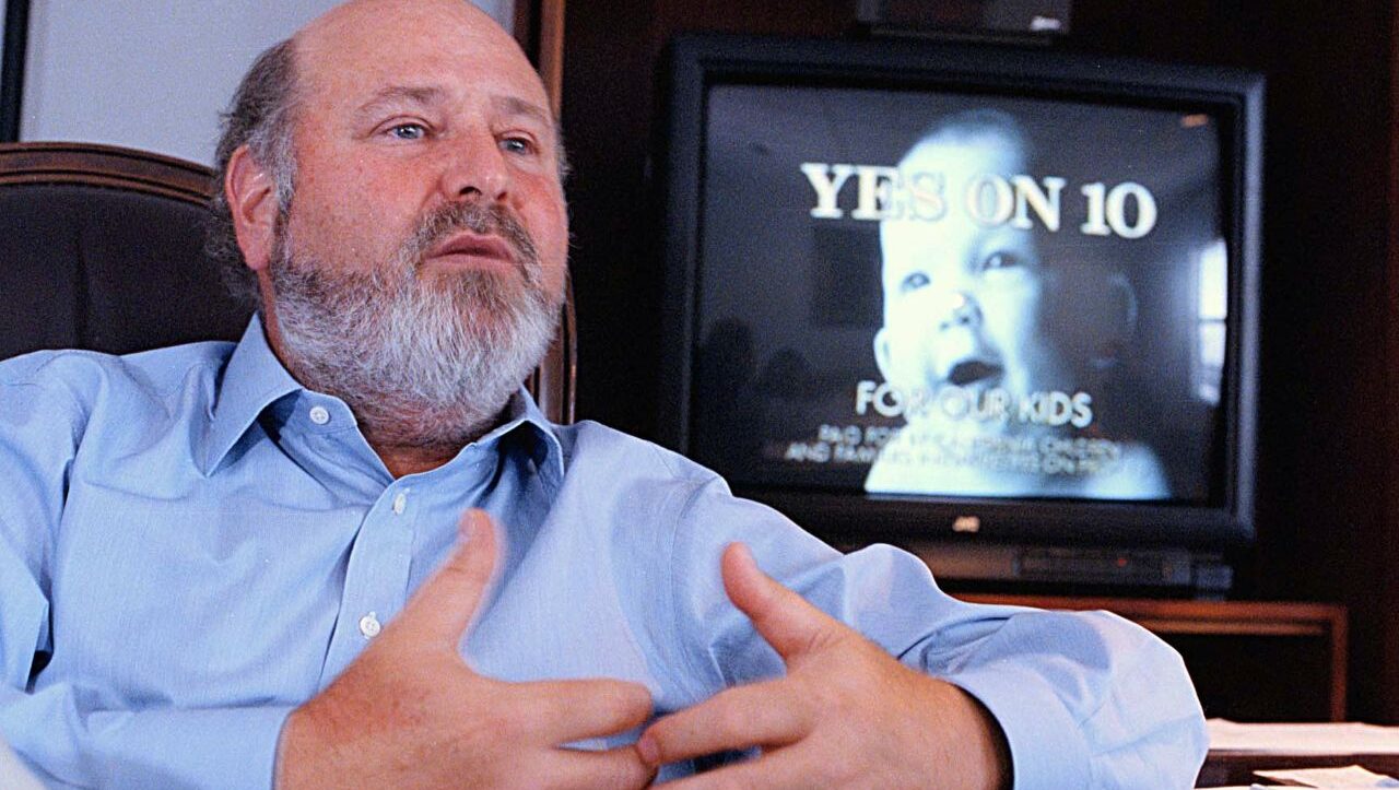 Image of Rob Reiner from 1998, sitting, wearing a blue shirt and talking about California's Proposition 10