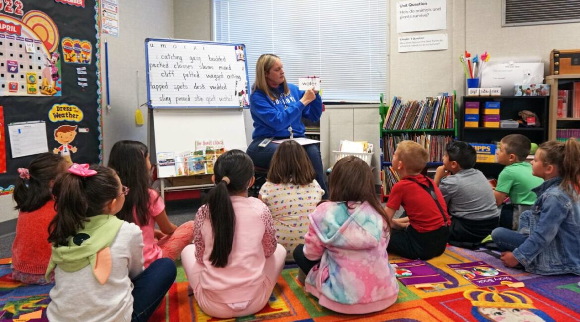 Image of a teacher in a blue sweatshirt teaching first-graders sitting on a quilt to read in a brightly decorated classroom
