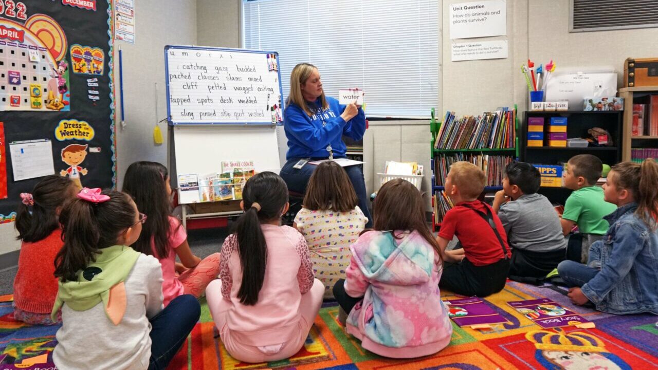 Image of a teacher in a blue sweatshirt teaching first-graders sitting on a quilt to read in a brightly decorated classroom
