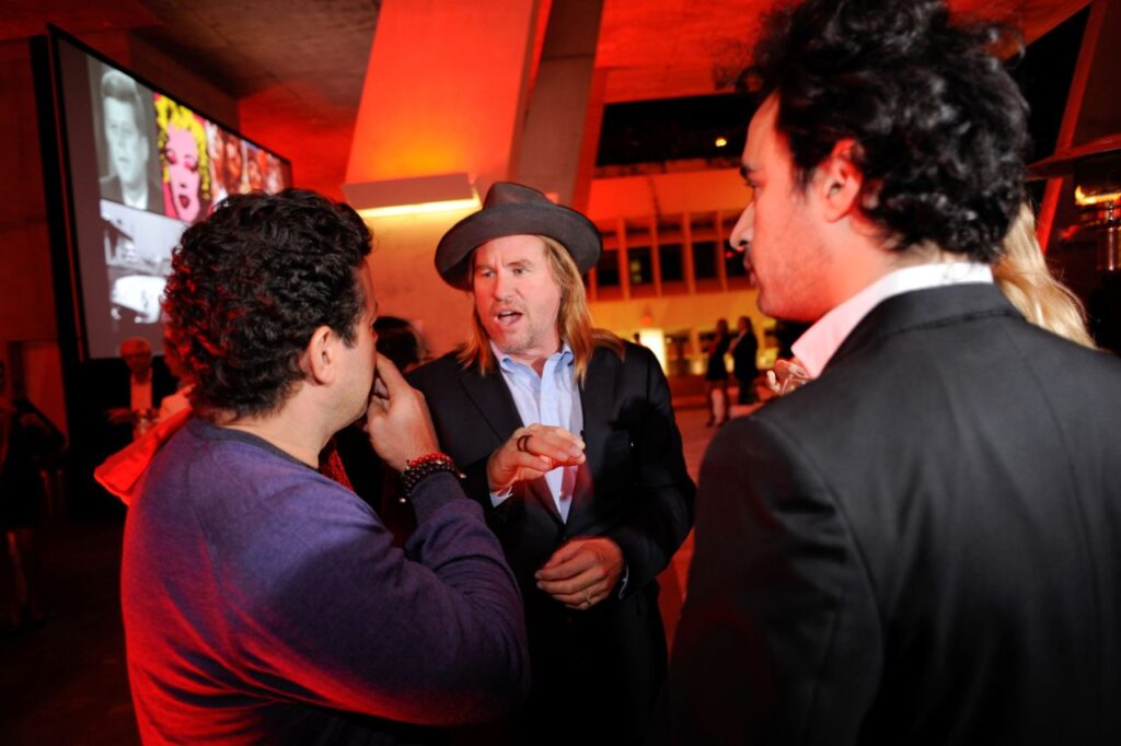 Image of actor Val Kilmer, with long blond hair and wearing a brown hat, talking to two men at a party