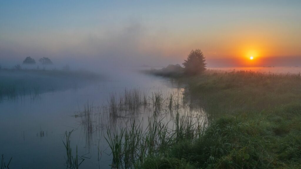 Image of the rising sun in a tableau that includes farm fields, fog, and a lake