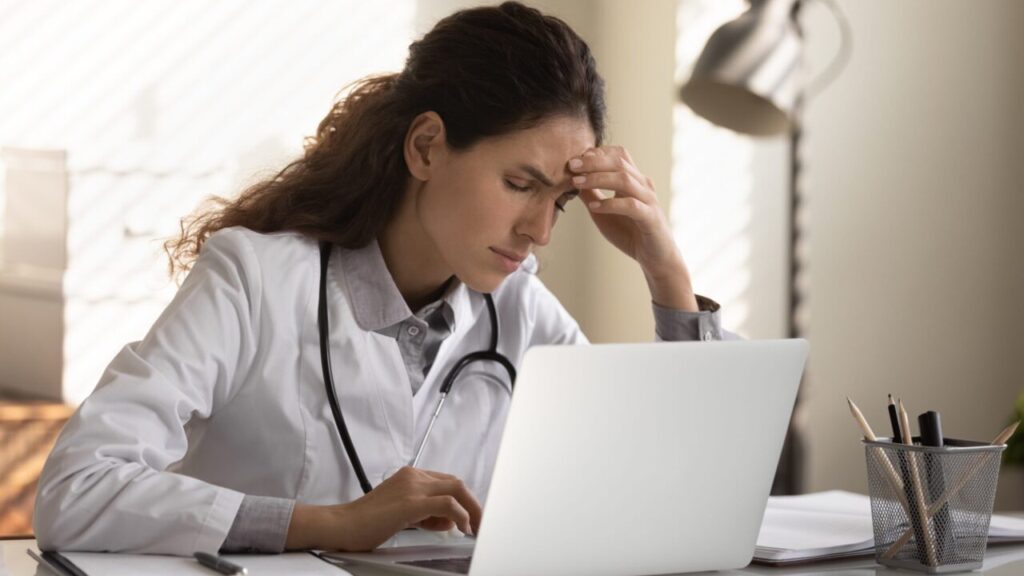 Image if a female physician sitting in front of a laptop with her hand on her forehead, suggesting fatigue or burnout