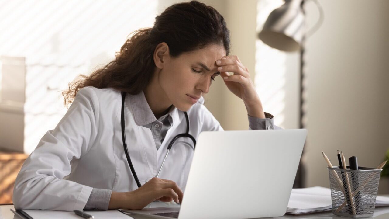 Image if a female physician sitting in front of a laptop with her hand on her forehead, suggesting fatigue or burnout