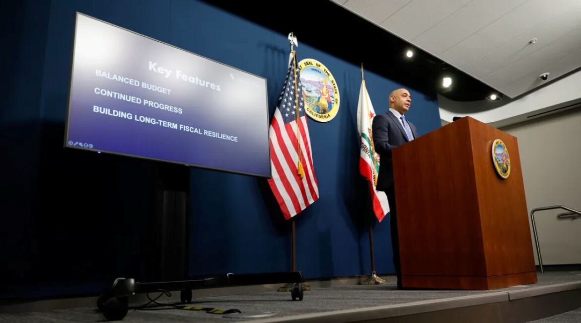 Image of California's budget director on stage unveiling the 2026-27 outlook. He's in a suit and flanked by the American and California flags