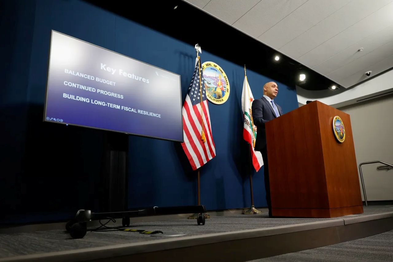 Image of California's budget director on stage unveiling the 2026-27 outlook. He's in a suit and flanked by the American and California flags
